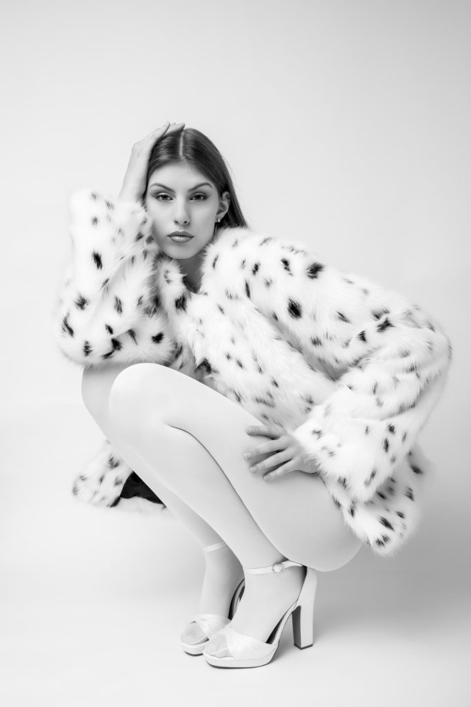 Portrait mode noir et blanc d’une femme accroupie en studio, manteau texturé clair, regard caméra